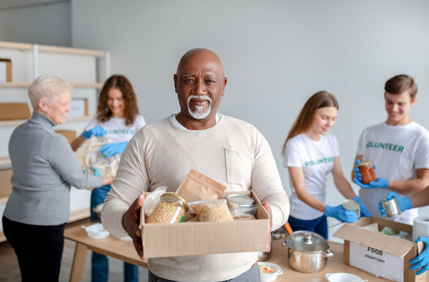 Charity center. Happy black senior man holding box with donations food, looking and smiling at camera. Group of volunteers giving elderly people boxes with products on background